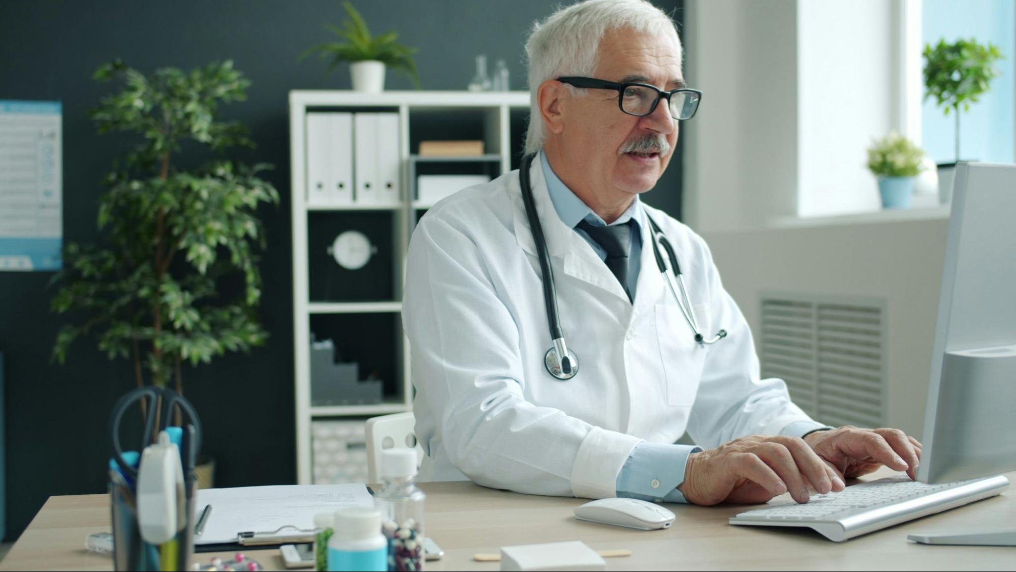A pharmacist with gray hair and glasses sits at a desk typing on a computer in a bright, modern office.