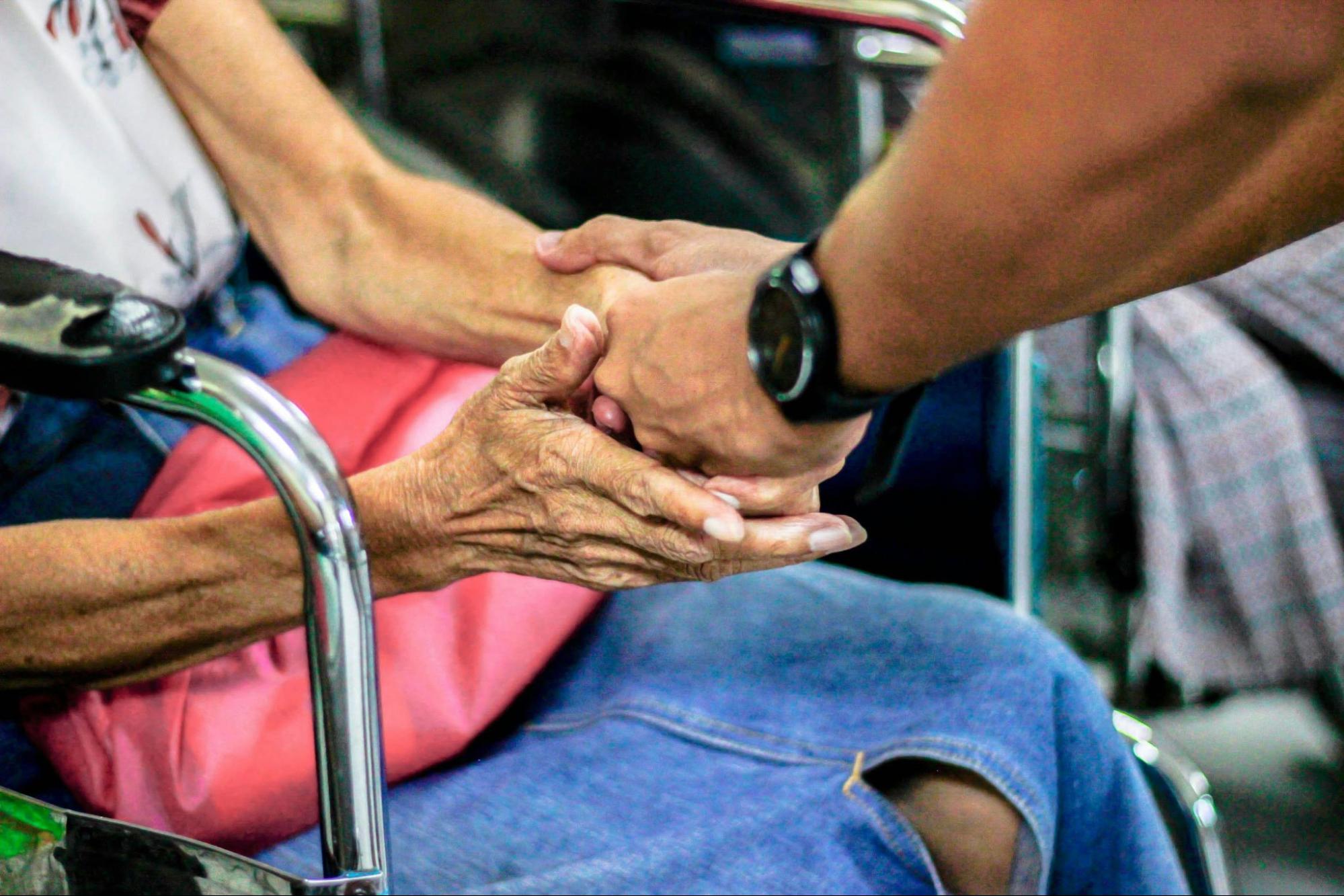 A caregiver gently holds the hands of an elderly person seated in a wheelchair, conveying support and compassion.