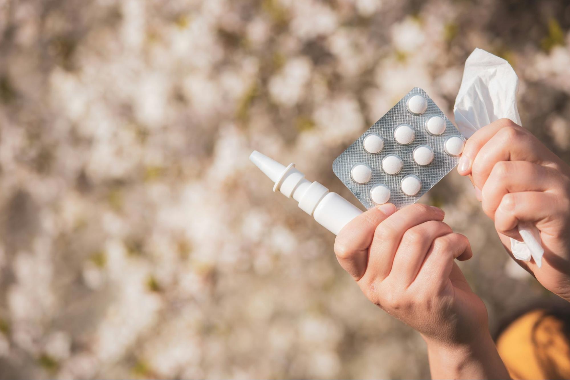 A person holds a nasal spray, a blister pack of tablets, and a tissue against a softly blurred outdoor background.