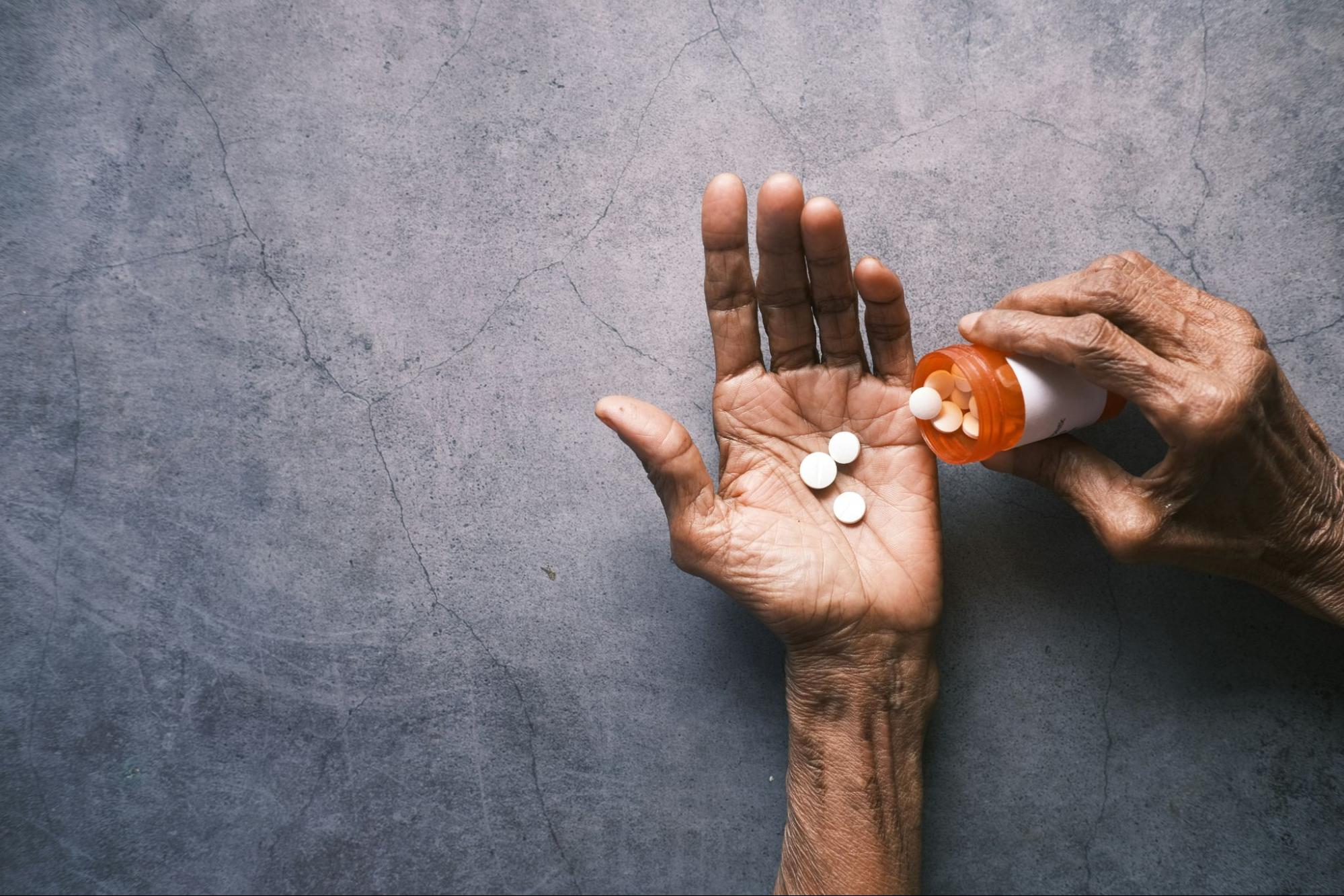 An older adult’s hand holds several white pills as another hand tips an orange prescription bottle over it against a gray surface.