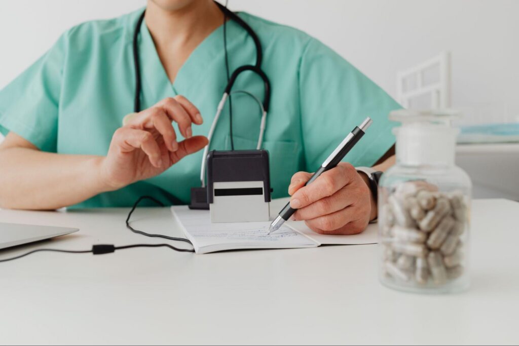 A healthcare professional wearing green scrubs and a stethoscope sits at a desk, writing in a notepad with a pen in one hand and preparing a self-inking stamp with the other, with an unlabeled jar of capsules in the front.