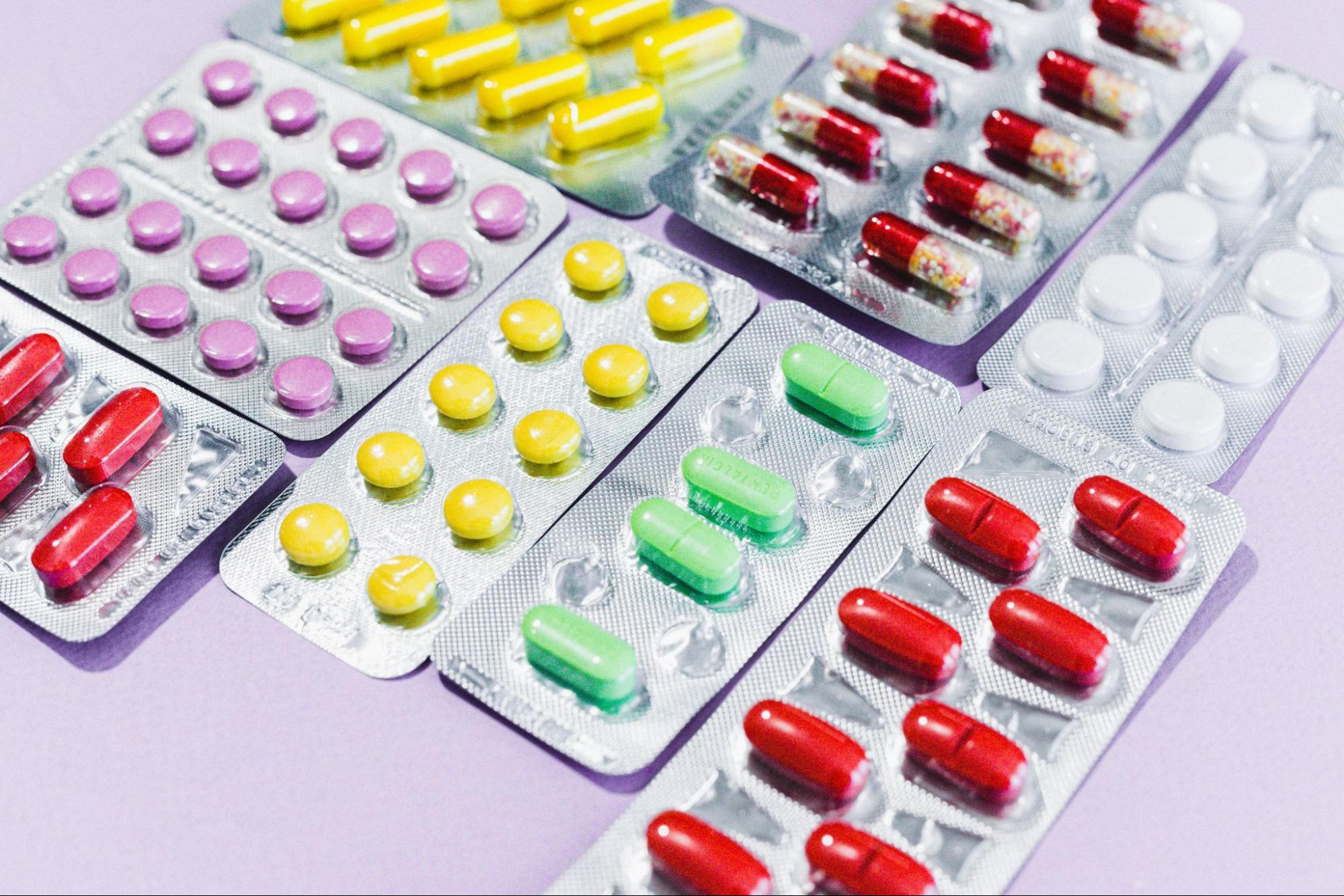 An assortment of multiple blister packs containing various colorful pills and capsules is arranged on a light purple surface.
