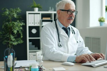 A pharmacist with gray hair and glasses sits at a desk typing on a computer in a bright, modern office.