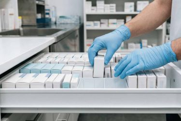 Close-up of gloved pharmacist’s hands arranging medication boxes in a white pharmacy drawer, no visible text or logos, contemporary medical environment, focus on order and cleanliness.